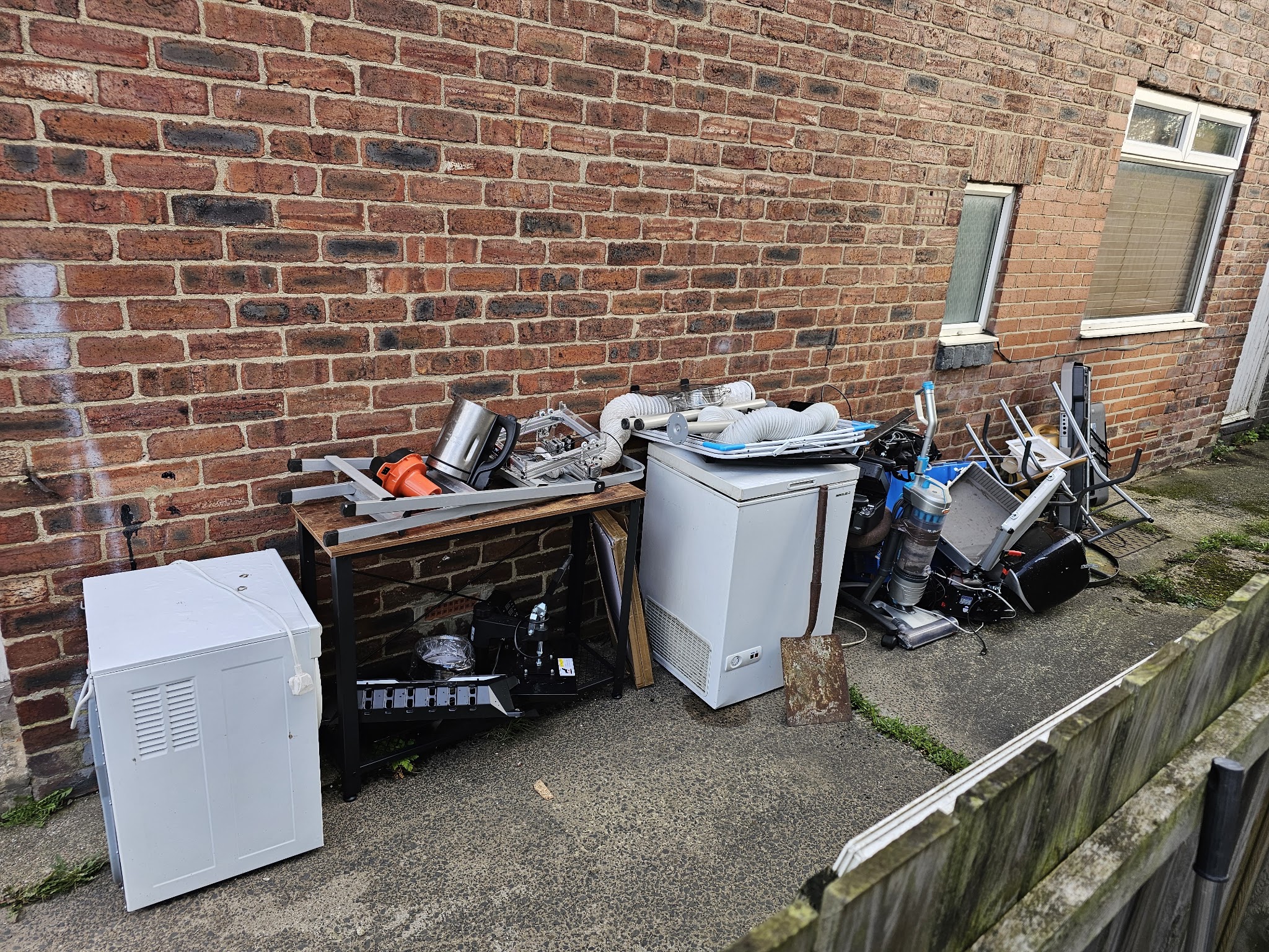 A chest freezer, mini fridge, table and household items stacked against a brick wall ready for collection